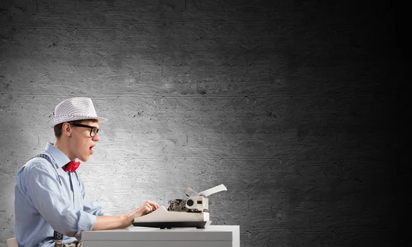 年轻震惊的男子作家在帽子和眼镜使用打字机 而坐在桌子上反对灰色混凝土墙的背景 — 图库照片
