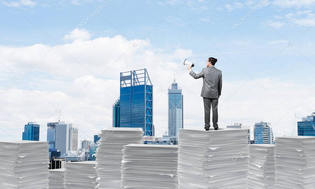 Businessman in suit standing on pile of documents with speaker in hand with cityscape on background. Mixed media.