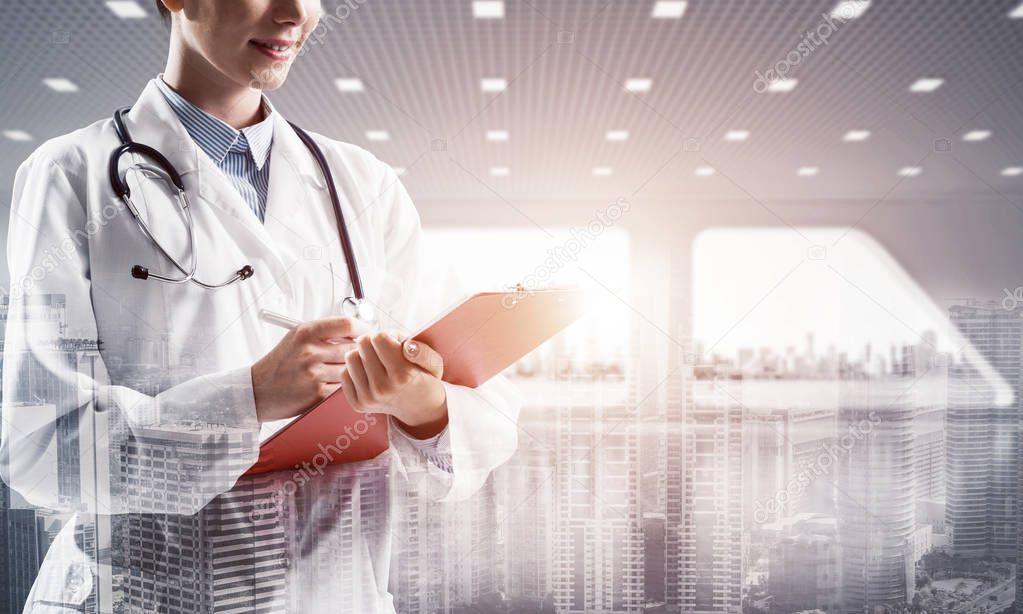 Side view of female doctor in sterile white coat making notes while standing at bright hospital office with modern cityscape view on background. Modern medical industry concept. Double exposure