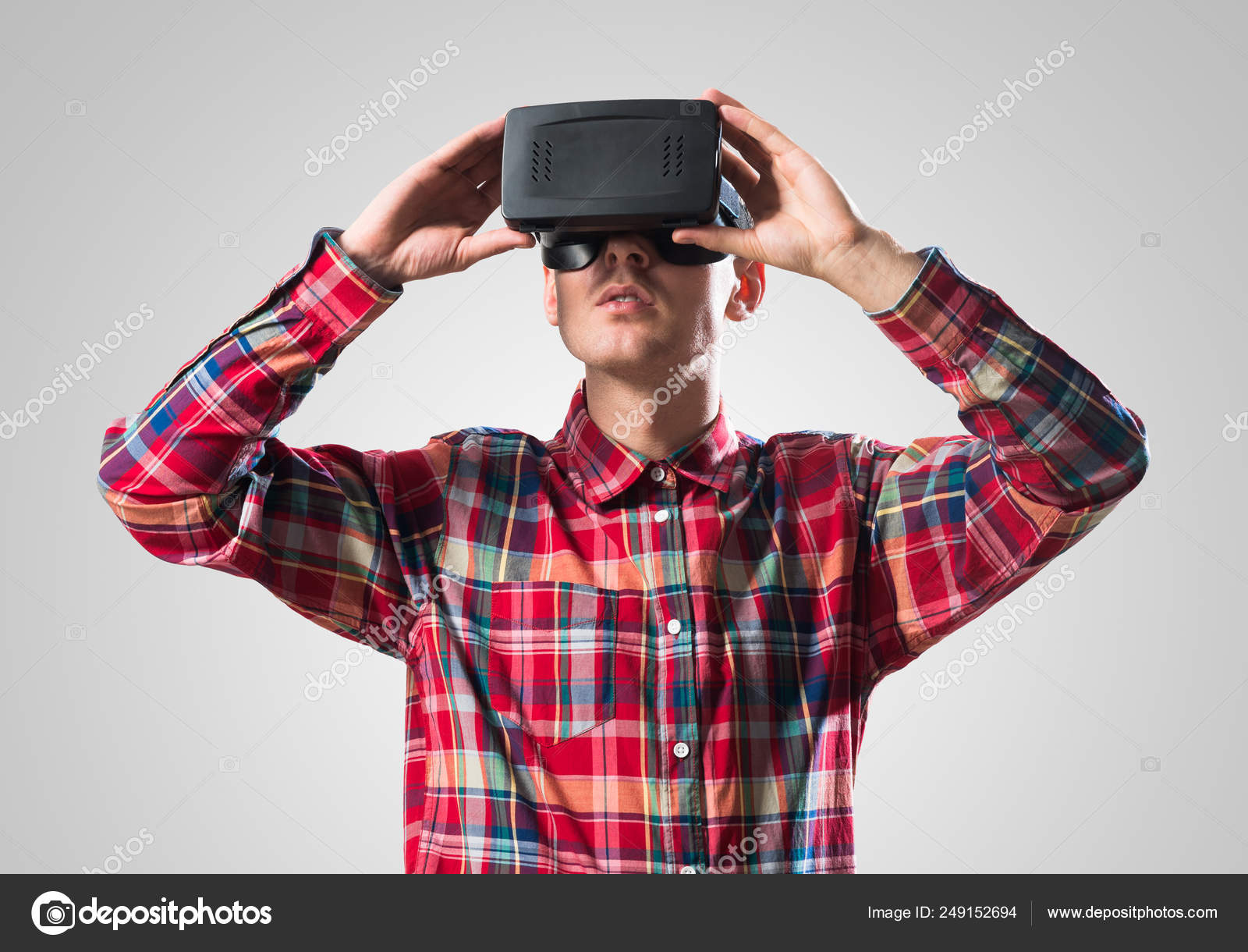 灰色の背景に対して仮想のヘルメットを身に着けているチェックのシャツの若い男 ストック写真 C Khakimullin