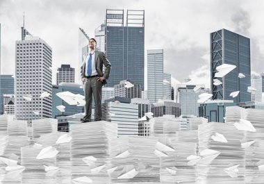 Takım elbiseli kendine güvenen işadamı arka planda şehir manzarası olan uçan kağıt uçaklar arasında evrak yığınının üzerinde duruyor. Karışık ortam.