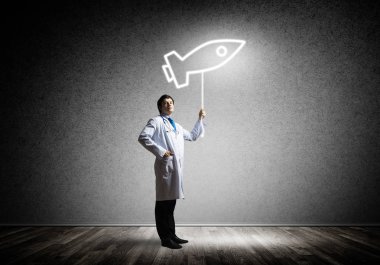 Bir arka plan üzerinde koyu gri duvarın karşısında dururken parlayan roket sembolü holding beyaz tıbbi üniformalı genç emin doktor yatay atış.