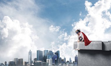 Maskeli ve pelerinli küçük üzgün çocuk binanın kenarında oturuyor ve aşağıya bakıyor. Karışık ortam