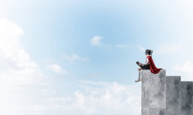 Maskeli ve pelerinli küçük çocuk beton bir grafikte oturup kitap okuyor. Karışık ortam