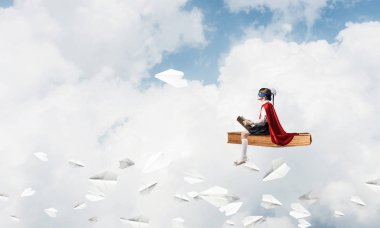 Maskeli ve pelerinli küçük çocuk gökyüzünde yüzüyor ve kitap okuyor.