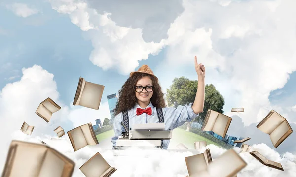 美丽的女作家在帽子和眼镜使用打字机和指向向上 而坐在桌子上的飞行书籍与浮动城市岛屿和多云的天空景背景 — 图库照片