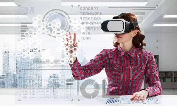 Educational process with help of VR headset. Cheerful young woman in checkered shirt using digital media interface while sitting indoors of office building.