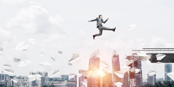 Businessman jumping over gap with flying paper planes in concrete bridge as symbol of overcoming challenges. Cityscape with sunlight on background. 3D rendering.