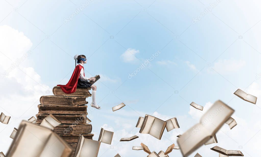 Little child in mask and cape sittting on pile of old books in sky and reading