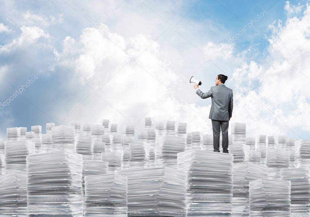 Businessman in suit standing on pile of documents with speaker in hand with cloudly skyscape on background. Mixed media.