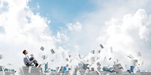 Businessman looking away while sitting on pile of documents among flying papers with cloudly sky on background. Mixed media.