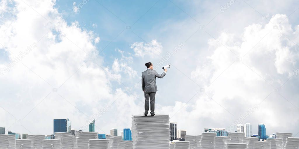 Businessman in suit standing on pile of documents with speaker in hand with skyscape and city view on background. Mixed media.