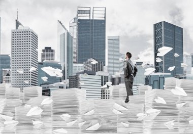 Takım elbiseli kendine güvenen işadamı arka planda şehir manzarası olan uçan kağıt uçaklar arasında evrak yığınının üzerinde duruyor. Karışık ortam.