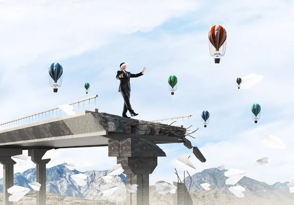 Businessman walking blindfolded among flying paper planes on concrete bridge with huge gap as symbol of hidden threats and risks. Flying balloons and nature view on background. 3D rendering.