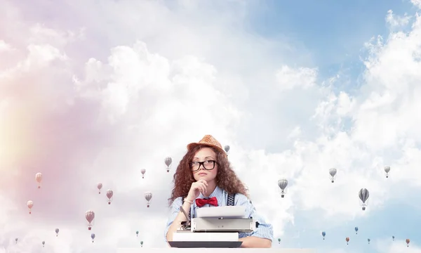 年轻和美丽的女作家在帽子和眼镜使用打字机 而坐在桌子上飞行轻飞行器和多云影像背景 — 图库照片
