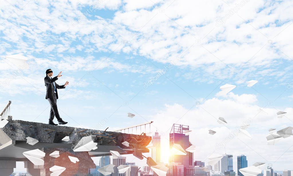 Businessman walking blindfolded among flying paper planes on concrete bridge with huge gap as symbol of hidden threats and risks. Cityscape and sunlight on background. 3D rendering.
