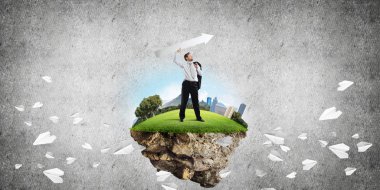 Takım elbiseli genç işadamının uçan kağıt uçakların arasında elinde büyük beyaz bir okla uçan adada dikilirken ve arka planda koyu gri bir duvara yaslanırken kavramsal görüntüsü..