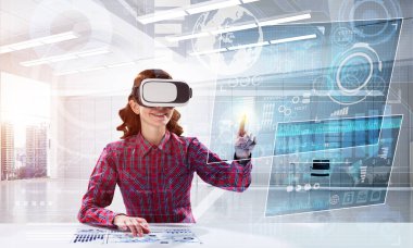 Parlak ofis binasında otururken dijital medya arayüzü olan VR kulaklık kullanan kareli gömlekli genç bir kadın. Eğitim için sanal gerçeklik teknolojileri
