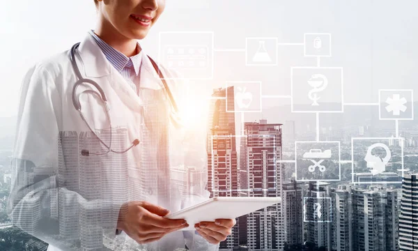 Cropped image of confident medical industry employee standing outdoors and holding tablet in hands. Young female doctor using tablet. Double exposure with medical interface icons