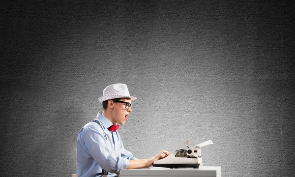 Young shocked man writer in hat and eyeglasses using typing machine while sitting at the table against gray concrete wall on background.