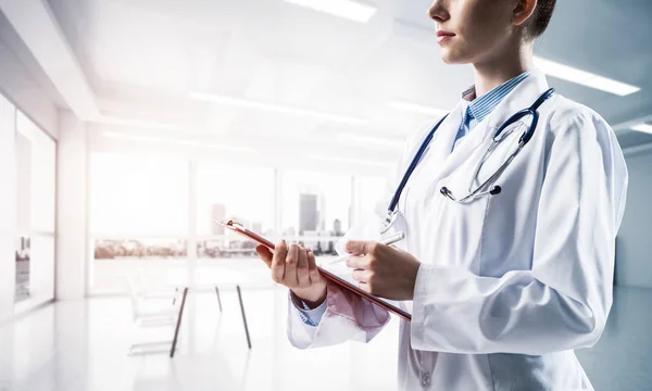 白い生殖不能のスーツの女性の医者の側面の眺めは背景に日光が付いている白い病院の建物の屋内に立っている間ノートに書く 医療業界のコンセプト — ストック写真