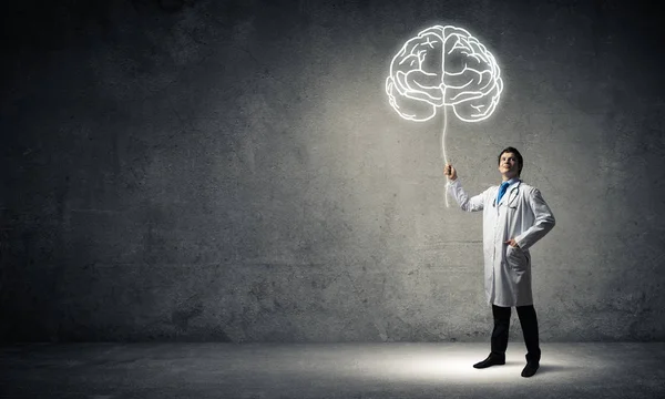 身着白色医服的年轻男医生站在空房间内 背景为灰色墙壁 与白色发光的大脑符号相交 — 图库照片