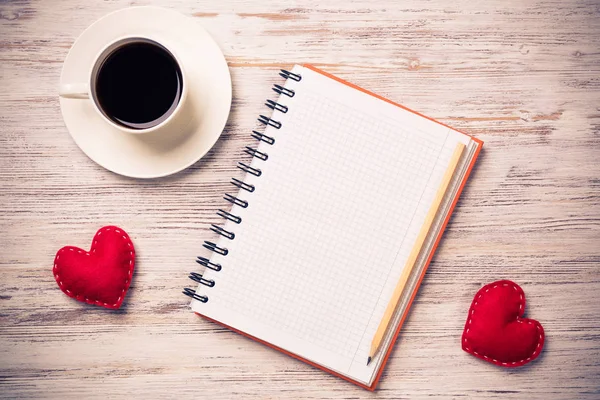 Coffee cup notepad pencil and two red hearts on wooden surface