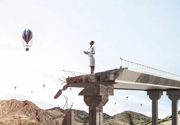 Confident medical industry employee in white uniform standing at the edge of broken bridge with cityscape view and flying aerostats on background. Medical industry concept