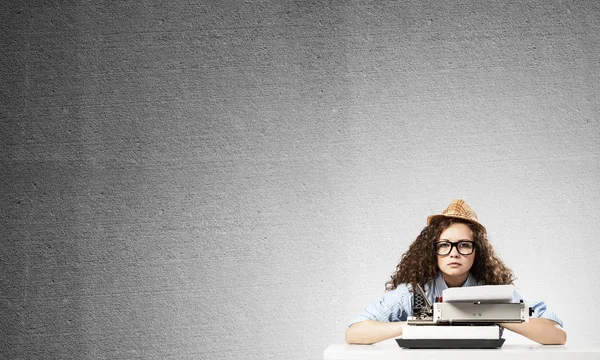 Young and beautiful woman writer in hat and eyeglasses using typing machine while sitting at the table against gray concrete wall on background.