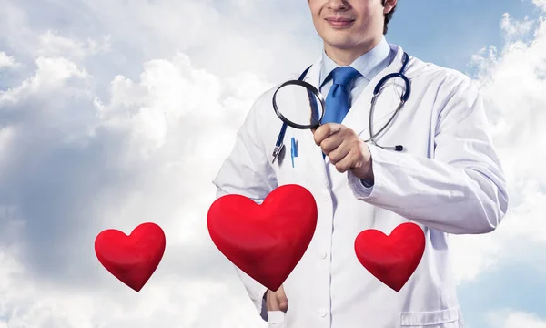 白い医療服を着た自信のある医師の水平ショットは 背景に曇ったスカイスケープビューに立っている間 拡大鏡を通して赤い心臓のシンボルを見ています — ストック写真