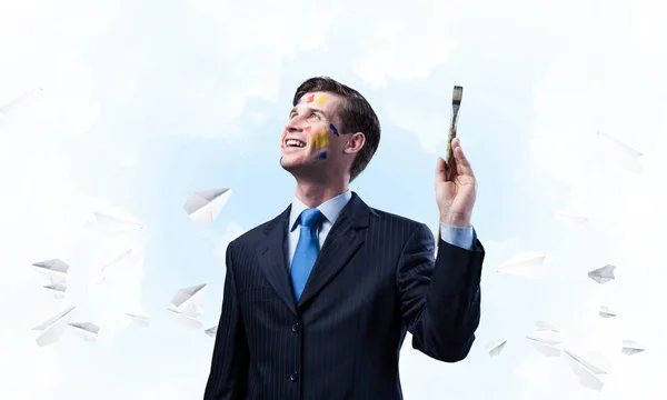 紙飛行機を背景に青い曇り Skyscape に対して立ったまま 彼の手に絵筆を持ち 微笑んでいる若いビジネスマンの概念的イメージ — ストック写真