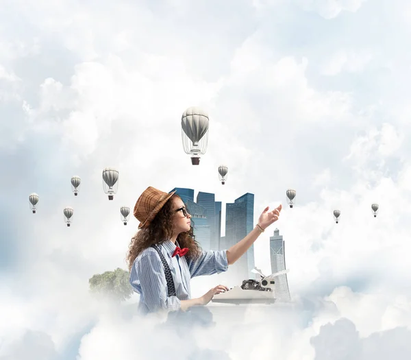 Young and beautiful woman writer in hat and eyeglasses using typing machine while sitting at the table with floating city island and cloudy skyscape with flying aerostats on background.