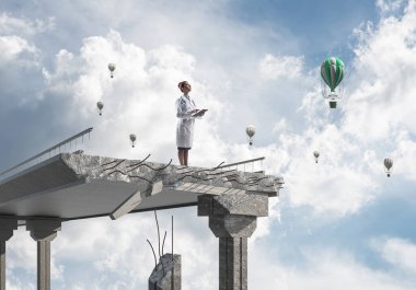 Peyzaj görünümü ve arka planda uçan hava balonları ile kırık köprünün kenarında ayakta iken tablet telefon kullanan profesyonel kadın doktor. Medikal Sanayi konsepti