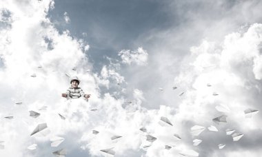 Küçük çocuk gözlerini kapalı tutuyor ve odaklanmış görünüyor. Arka planda bulutlu bir gökyüzü ile havada uçan kağıt uçaklar arasında meditasyon yapıyor..