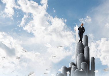 Takım elbiseli hırslı ve genç işadamı, arka planda bulutlu gökyüzü manzarası olan stune sütununda dururken elinde büyük beyaz bir afiş tutuyor.