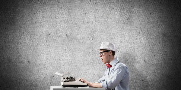 年轻震惊的男子作家在帽子和眼镜使用打字机 而坐在桌子上反对灰色混凝土墙的背景 — 图库照片