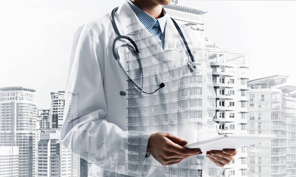 Double exposure of conceptual image of young female doctor standing with tablet in hands and modern city view on background. Medical industry