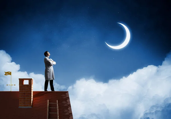 背景での夜の天空とレンガ屋根の上部に白い医療制服立っている若い医者の水平方向のショットの表示します — ストック写真