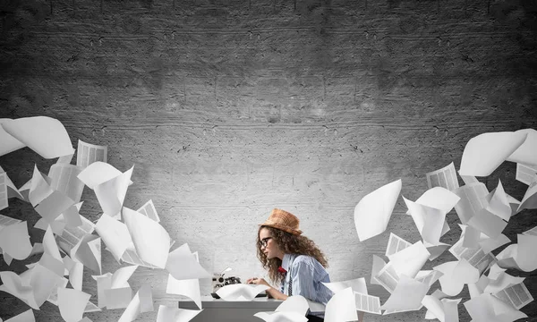 Young and beautiful woman writer in hat and eyeglasses using typing machine while sitting at the table among flying papers and against gray concrete wall on background.