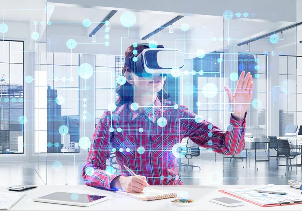 Horizontal shot of young woman in checkered shirt using virtual reality headset with media interface while sitting inside bright building. VR technologies for educational process