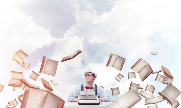 Young man writer in hat and eyeglasses using typing machine while sitting at the table among flying books with cloudy skyscape on background.