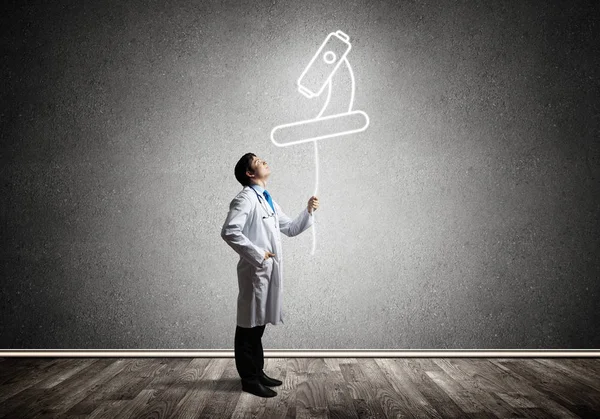 Arka planda gri duvara ayakta iken beyaz üniforma havada parlayan mikroskop simgesi ile interracting genç erkek doktorun kavramsal görüntü.