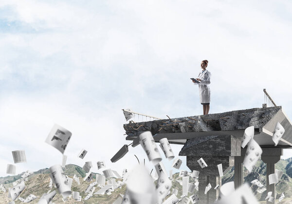 Young woman doctor in white medical uniform standing among flying papers on the edge of broken bridge with landscape view on background. Medical industry concept