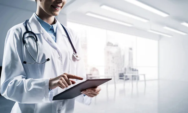 Cropped image of young woman doctor touching tablet screen with finger while standing inside bright medical office building with sunlight on background. Medical industry concept