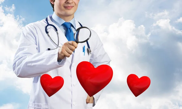 白い医療服を着た自信のある医師の水平ショットは 背景に曇ったスカイスケープビューに立っている間 拡大鏡を通して赤い心臓のシンボルを見ています — ストック写真