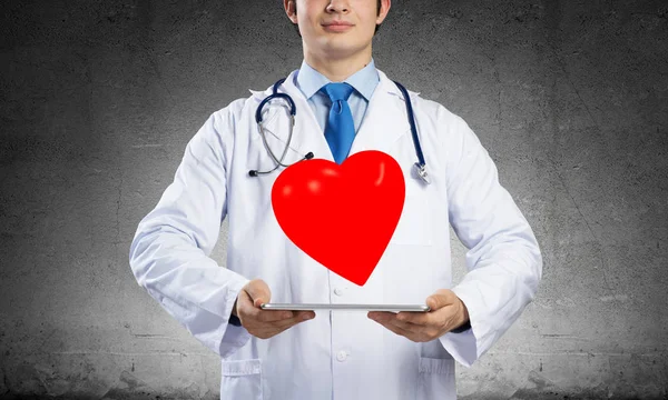 白い医療コートを着た陽気な若い男性医師の水平ショットは 灰色の背景に立っている間 赤いハートのシンボルを提示しています — ストック写真