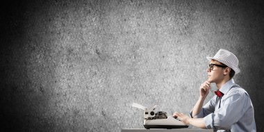 Genç yazar şapka ve gözlük arka planda gri beton duvara karşı masada otururken yazarak makinasıyla adam.