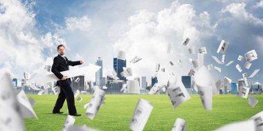 Takım elbiseli ve genç işadamı elinde büyük beyaz bir ok tutarak yeşil çimenlikte uçan kağıtların ve arka plandaki şehir manzarasının arasında duruyor..