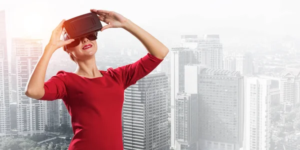 Conceptual image of young woman in red dress using virtual reality headset while standing outdoors with modern cityscape view on background
