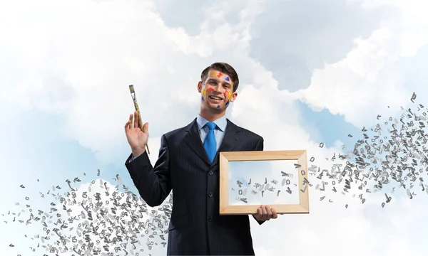 年轻和成功的商人的肖像 黑色西装举行相框和画笔在手 而站着与飞行字母反对蓝色多云的天空景观视图在背景 — 图库照片